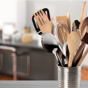 A metal container holds assorted kitchen utensils on a countertop, with a blurred kitchen background featuring chairs and shelves.