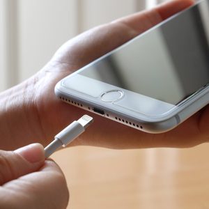 A hand holds a smartphone while inserting a charger cable into the port, with a blurred indoor background.