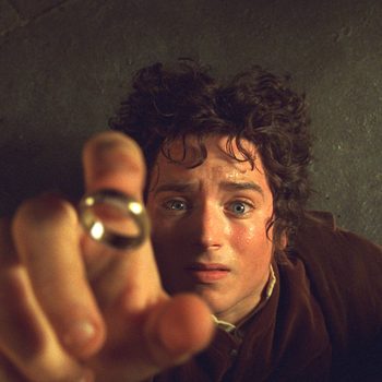 A person holds a ring with an outstretched hand, looking intently upwards, set against a dim, stony background.