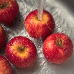 Red apples being washed under running water in a stainless steel sink.