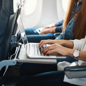 Laptop sits on an airplane tray table, being used by a person typing, surrounded by other passengers seated in a row.