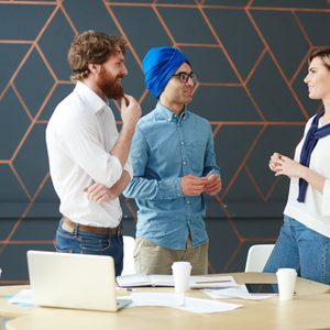 Three people standing, engaged in conversation, around a modern office table with laptops and papers, against a geometric-patterned wall backdrop.
