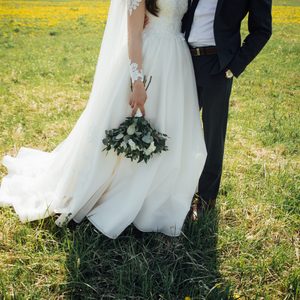 Bride holding bouquet stands with groom in a grassy field, surrounded by yellow flowers, under a sunny sky.