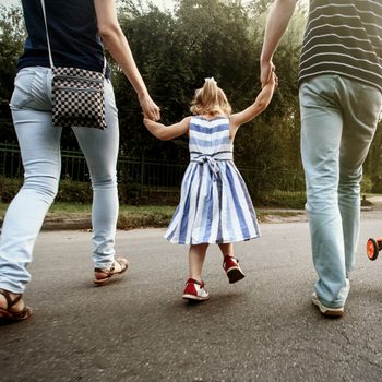 A child, holding hands with adults, walks on a road, with a scooter alongside. Trees and grass line the path.