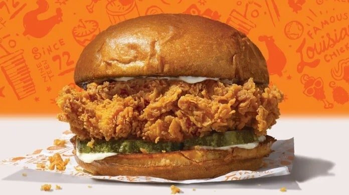 A crispy fried chicken sandwich sits on a wrapper, featuring pickles and mayonnaise, with an orange background covered in illustrated text and graphics.