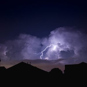 Lightning illuminates dark storm clouds, striking across a night sky above silhouetted rooftops and trees.