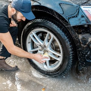 Person cleans car wheel with a sponge, surrounded by soapy water on a concrete surface.