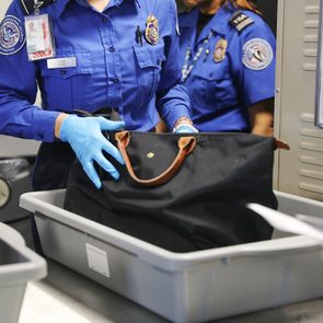 TSA officer inspects black bag in a tray at airport security checkpoint while wearing blue uniform and gloves.