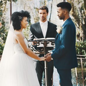 Bride and groom hold hands under floral arch during outdoor wedding ceremony, officiated by a man in a suit.