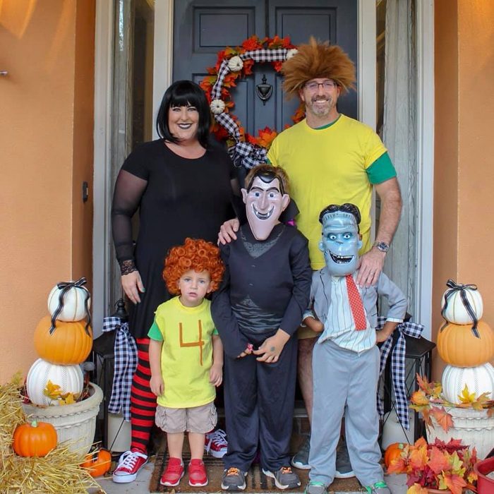 Family poses in Halloween costumes on a decorated porch with pumpkins and autumn wreath, one child wearing a shirt with the number 