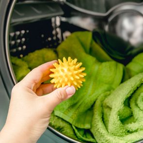 A hand holds a yellow spiky laundry ball, placing it in a washing machine with green towels inside.
