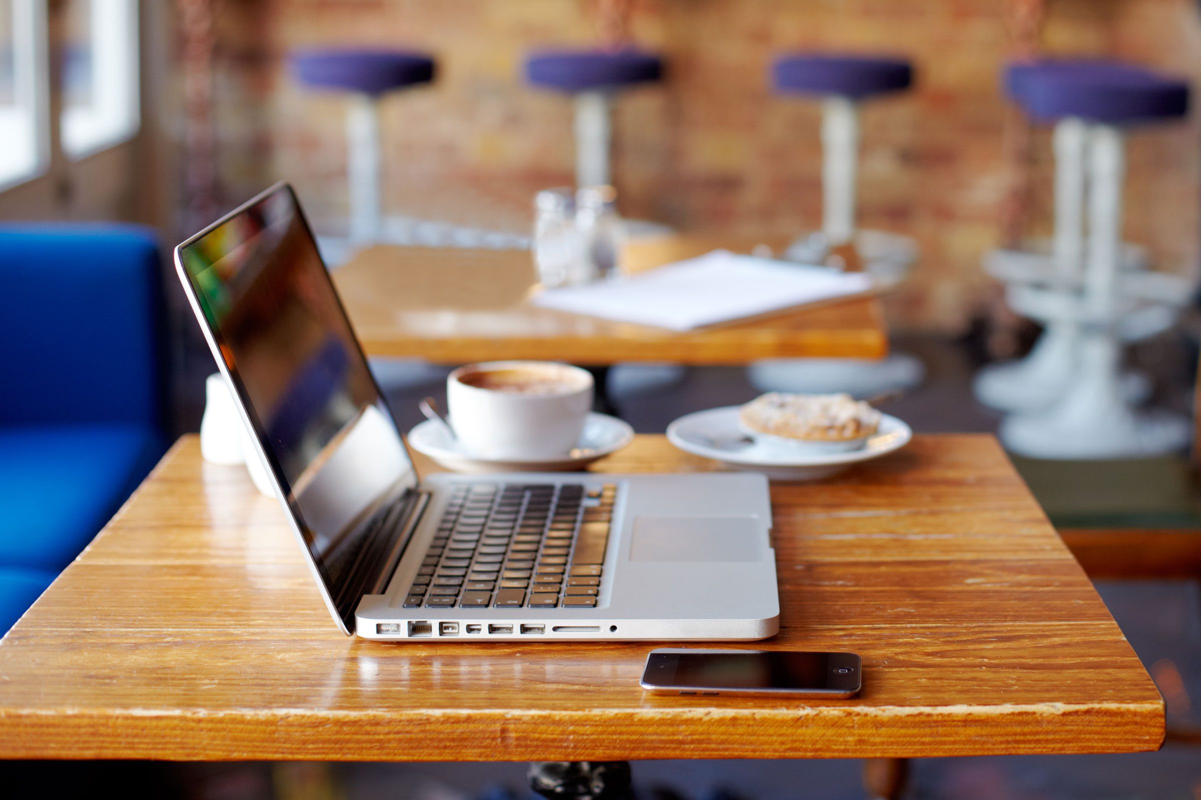 Laptop resting on a wooden table, accompanied by a smartphone and coffee, in a cozy cafÃ© with blue seats and blurred stools.