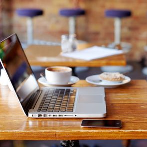 Laptop resting on a wooden table, accompanied by a smartphone and coffee, in a cozy cafÃ© with blue seats and blurred stools.