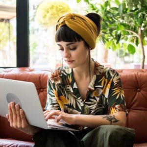 Person typing on a laptop, sitting on a brown leather couch, surrounded by lush green plants near a large window.