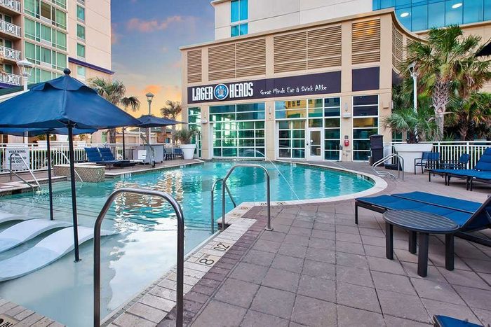 The pool glistens under umbrellas, surrounded by lounge chairs, with a building labeled 