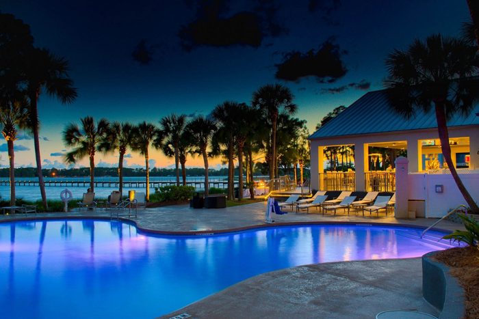 A glowing pool reflects purple lighting, surrounded by palm trees and lounge chairs at sunset, with a waterfront view and illuminated building nearby.