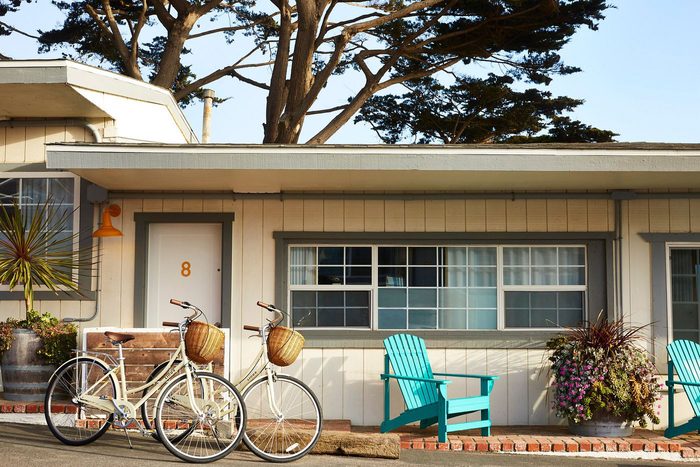 Bicycles rest outside a motel room, flanked by blue chairs and flowerpots, under trees in a sunny outdoor setting.