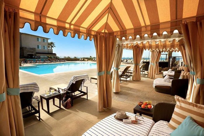 Lounge chairs rest under cabanas beside a pool, amid a resort setting with palm trees and buildings in the background.