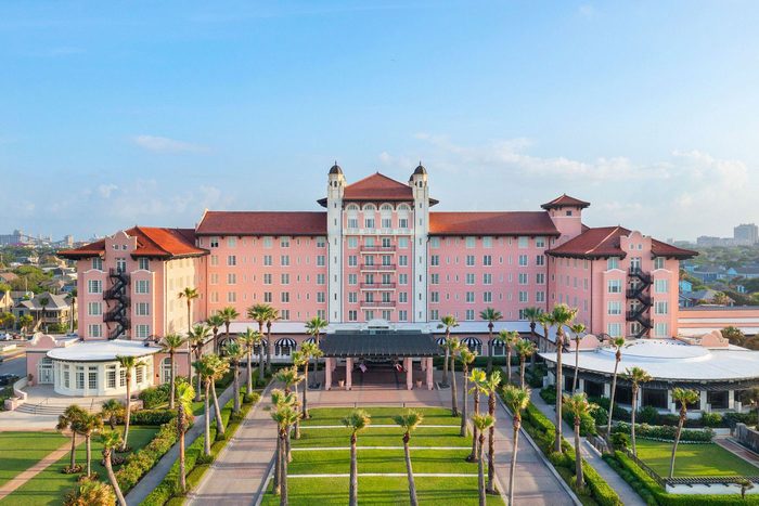 Pink hotel stands surrounded by palm trees, featuring a red roof and lush gardens, under a clear blue sky.