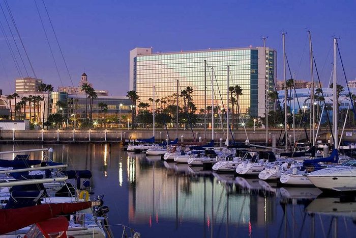 Boats rest docked on calm water, reflecting city lights; a large glass building and palm trees line the waterfront at dusk.