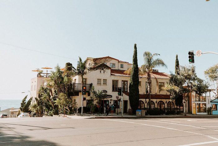 A Spanish-style hotel sits on a sunny street corner, surrounded by palm trees. People walk near the entrance, and traffic lights are visible.