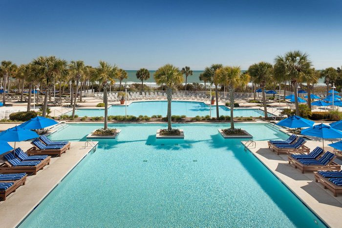 Pool, shimmering under sunlight, surrounded by palm trees and blue-striped loungers. Blue umbrellas provide shade, with the ocean visible in the background.