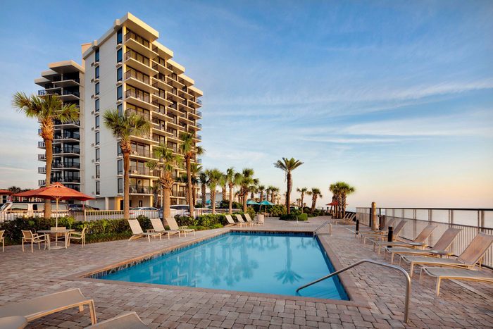 A swimming pool sits surrounded by lounge chairs, near a tall building, with palm trees and an ocean view under a clear sky.