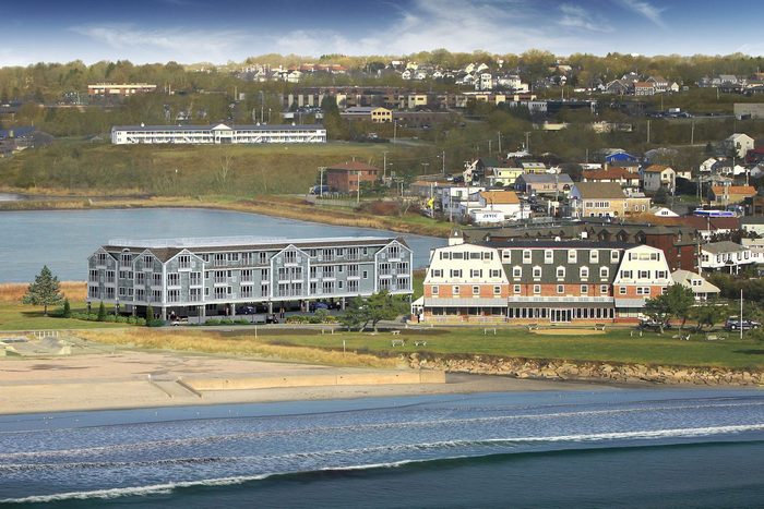 Buildings stand beside a beach, framed by a grassy area, and a town rests on distant rolling hills under a partly cloudy sky.