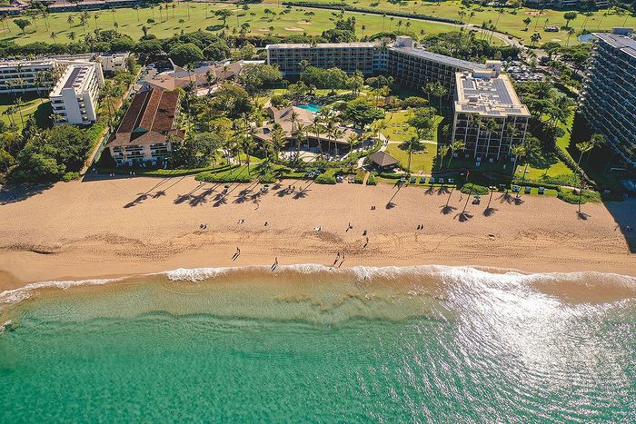 Beachfront resorts overlook ocean waves as people walk along the sandy shore, surrounded by lush greenery and golf course in the background.