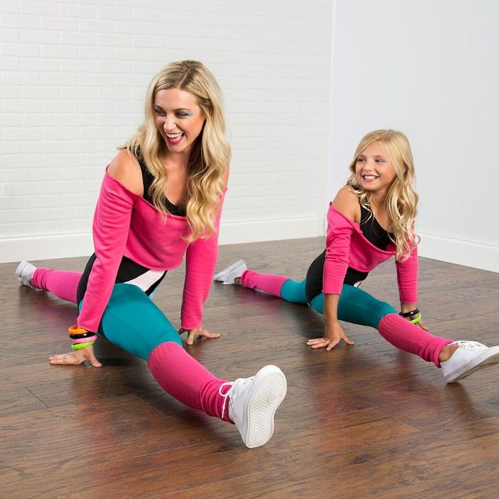 Two individuals performing splits on a wooden floor, wearing colorful workout outfits, smiling in a bright room with white brick walls.