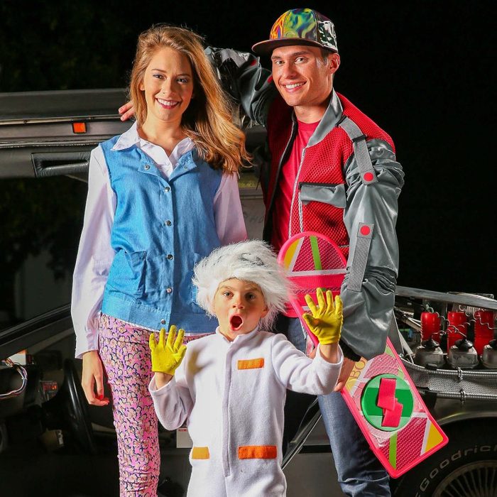 Three people wearing colorful costumes smile in front of an open car door, with a child raising hands and a hoverboard visible.