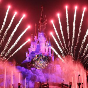 Fireworks explode above an illuminated castle, surrounded by colorful fountains at night in a festive setting.