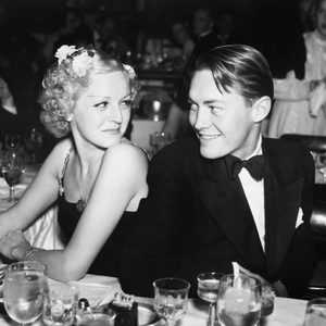 A man and woman sit smiling at each other in a formal dining setting, surrounded by elegantly dressed people and tableware.