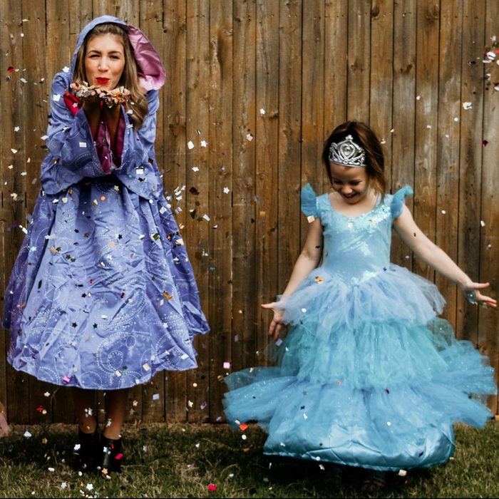 Two people in costumes joyfully throw colorful confetti, standing on grass against a wooden fence backdrop.