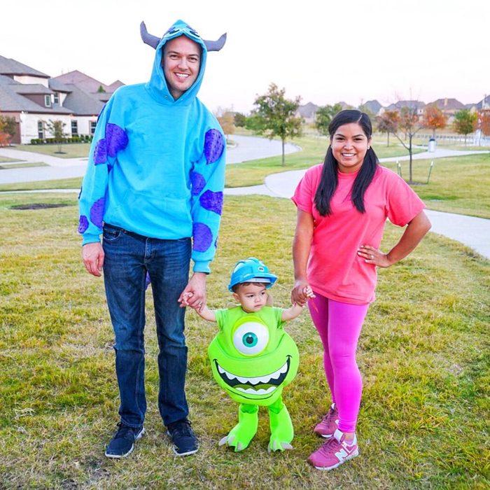 Three people stand holding hands; one in a blue hoodie with horns, another in a green monster costume, and one in pink, on grass.