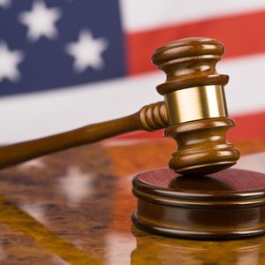 A wooden gavel rests on a sound block, set against a blurred backdrop of the American flag's stars and stripes.