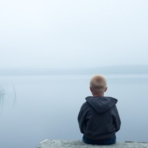 A child sits quietly, gazing at a serene, misty lake with reeds to the left.