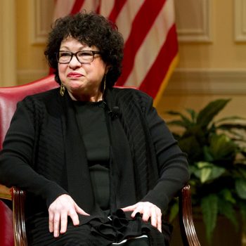 A person sits in a red chair, smiling slightly, in a formal setting with an American flag and plants in the background.