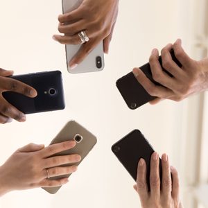 Five hands holding smartphones, reaching inward, creating a circle-like formation against a bright, neutral background.