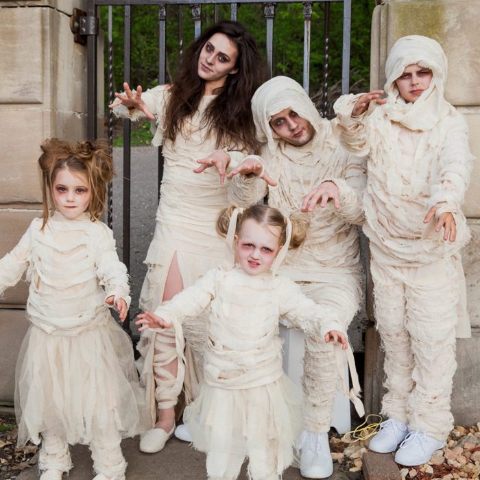 People dressed as mummies pose playfully, acting spooky in front of a metal gate and stone pillars outdoors.