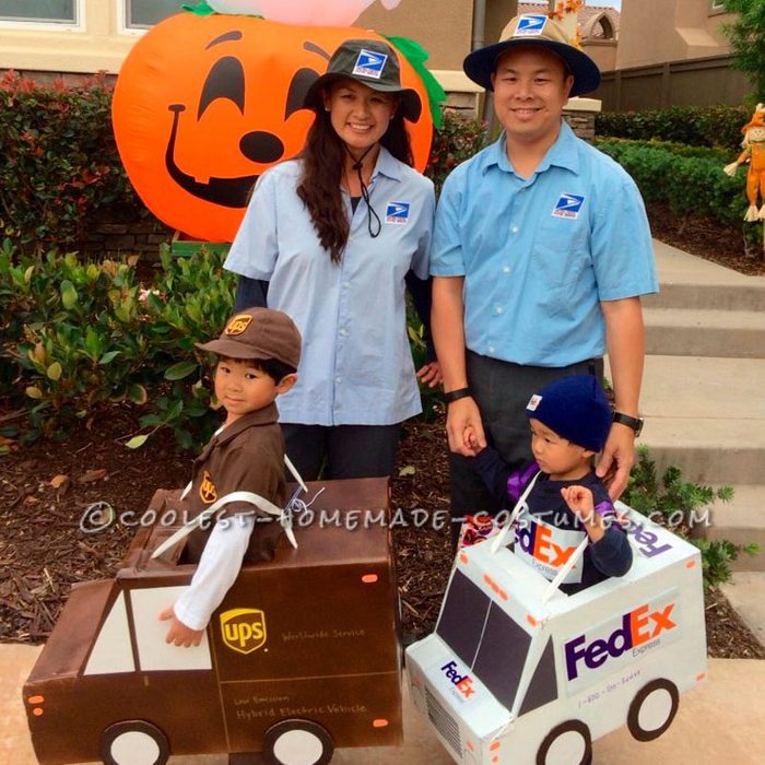 Two adults wear postal uniforms, smiling with two children dressed in UPS and FedEx cardboard vehicle costumes, standing outside by a large pumpkin decoration.
