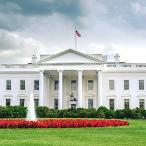 White house stands with columns, surrounded by trees and shrubs; a fountain and red flowers decorate the green lawn. Overcast sky above.