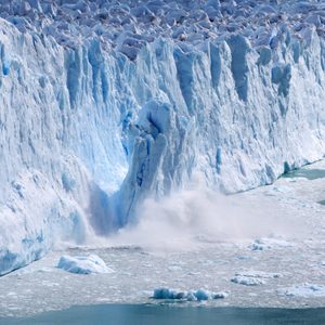A large glacier is calving, releasing chunks of ice into a cold, turquoise sea amid icy surroundings.