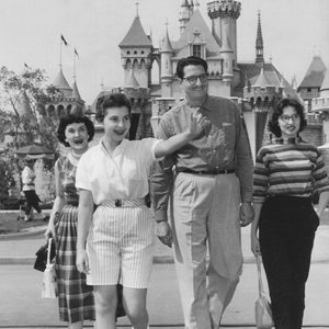 A group of four people walk happily in front of a castle, likely at a theme park, in a vintage photograph.