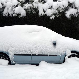 Car is buried under thick snow, parked beside snow-covered bushes.