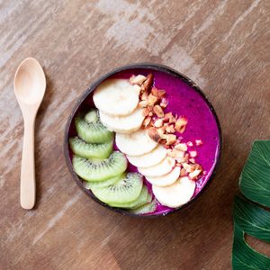 A smoothie bowl topped with kiwi, banana, and nuts sits on a wooden table next to a wooden spoon and a green leaf.
