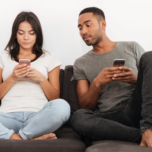Two people sit on a couch, using their smartphones, focused and engaged. The setting is relaxed and informal.