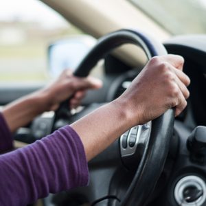Hands gripping steering wheel, driving on a road; interior of a car is visible, with blurred scenery outside.