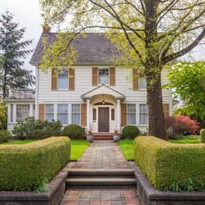 A two-story white house stands serenely, bordered by hedges and trees, with a cobblestone path leading to its front door in a lush garden.