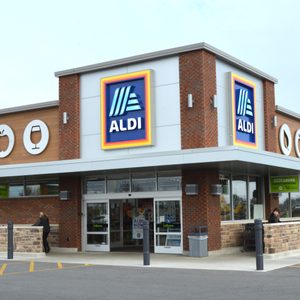 Aldi storefront displaying logo; customers enter. Red brick walls and grocery-themed icons highlight supermarket entrance under a cloudy sky.